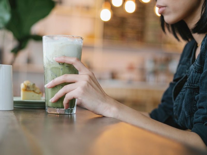 A hand around a glass of iced matcha latte at a cafe counter with warm string lights, original