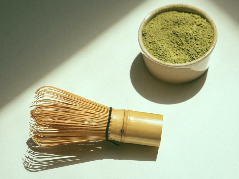 A bamboo matcha whisk and a small bowl of matcha powder on a white table in hard sunlight, with the matcha filter