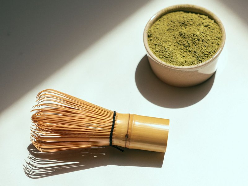 A bamboo matcha whisk and a small bowl of matcha powder on a white table in hard sunlight, original